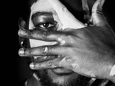Image of Black man with white paint on face, covering face with his hand, black and white